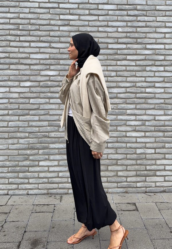 Woman wearing a black hijab, checked jacket, black wide pants, and brown sandals walking beside a gray brick wall.