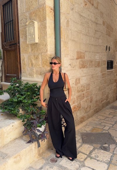 Top negro tipo halter y pantalones anchos negros con bolsillos frontales. Sandalias negras simples. Fondo de pared de piedra con plantas verdes.