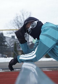 Athlète portant une veste de sport turquoise avec des bandes blanches, des gants noirs, des lunettes de soleil et une casquette noire, appuyé sur une rampe. Arrière-plan enneigé.