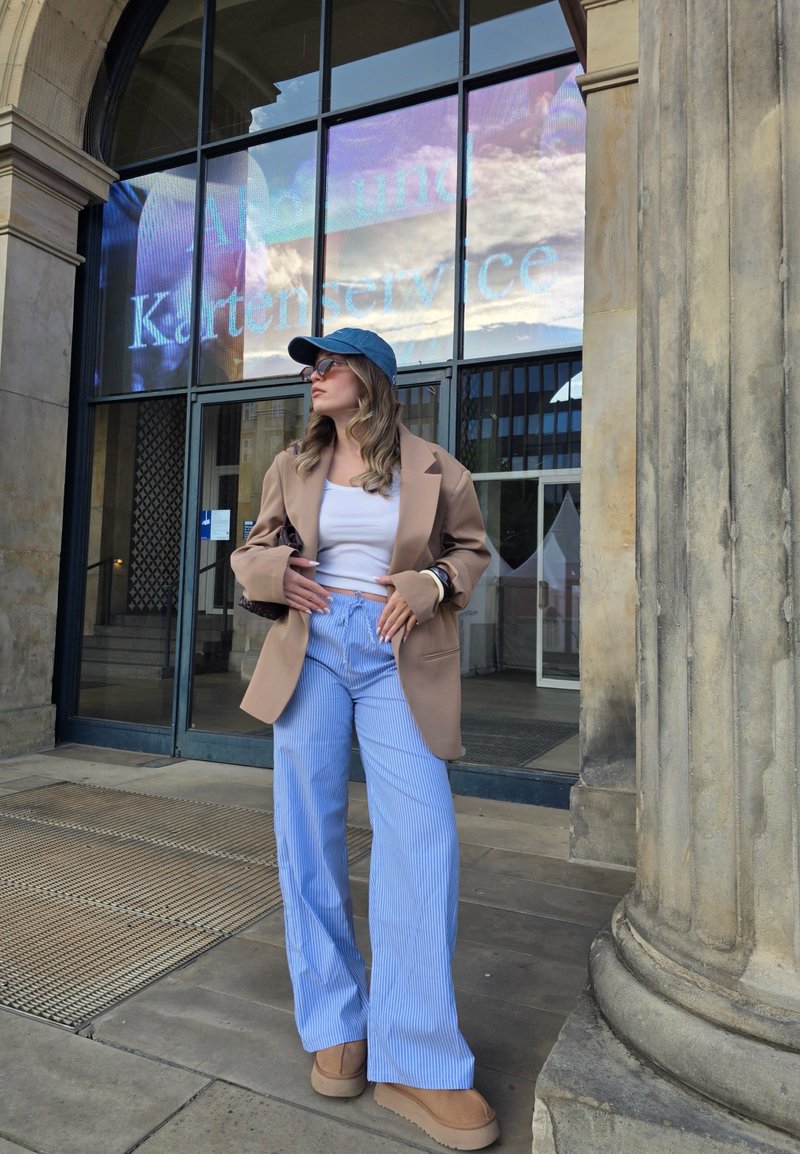 Blazer oversize beige, crop top blanc, pantalon large rayé bleu, chaussures compensées beiges et casquette bleue, se tenant devant l'entrée d'un bâtiment.