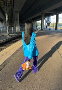 Blazer de cuero azul sobre pantalones de chándal morados, sosteniendo un juguete de peluche naranja en un bolso de cadena, caminando por un andén de tren bajo un paso elevado.