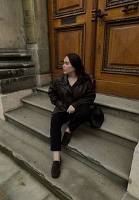 Braune Lederjacke, schwarze Hose und dunkle Schuhe, die auf steinernen Stufen neben einer Holztür sitzen. Eine dunkle Tasche daneben platziert. Glatte Texturen und gedeckte Farben.