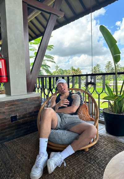 Silla de mimbre redonda con cojín gris, persona vestida con camiseta sin mangas negra y pantalones cortos, zapatillas blancas y una gorra en un entorno exterior con muchas hojas.