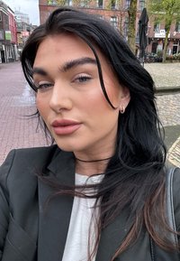 Primo piano di una persona con lunghi capelli neri, che indossa un blazer nero sopra una maglietta bianca, trucco minimal e orecchini a cerchio piccoli.