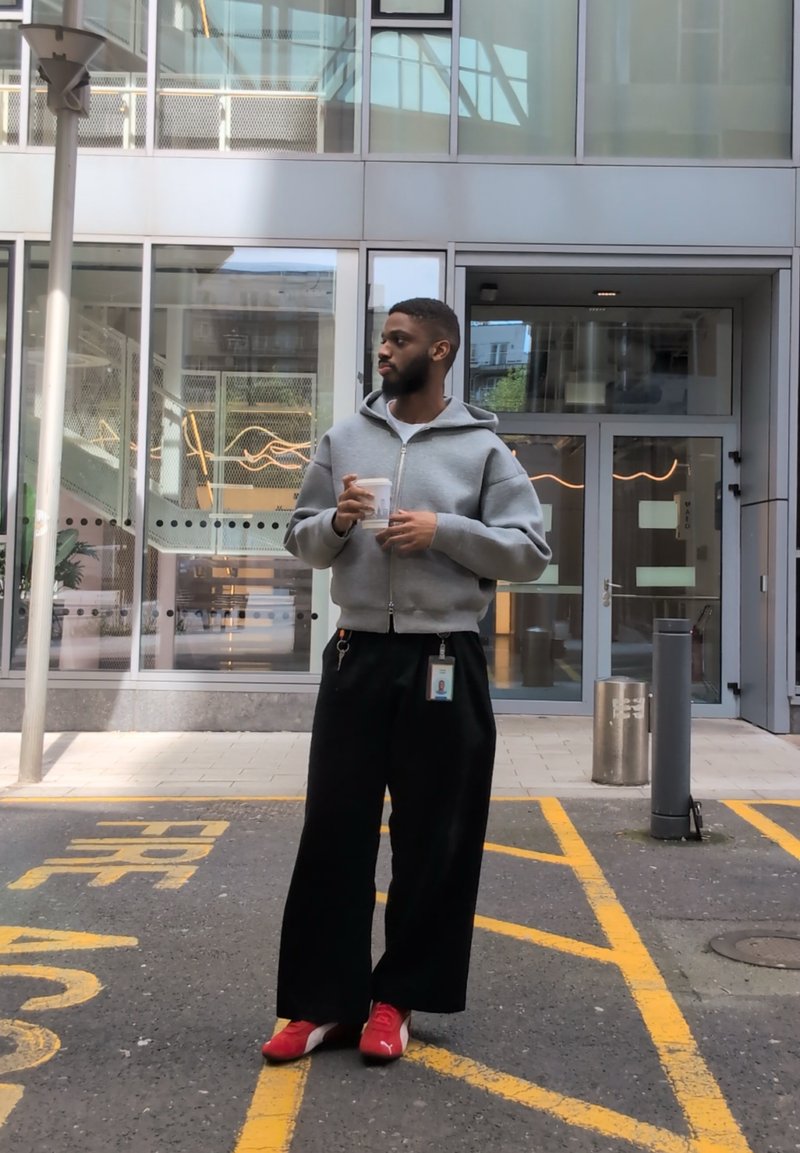Homme en sweat à capuche gris et pantalon noir tenant une tasse, debout devant un bâtiment moderne en verre dans une zone de trottoir marquée.