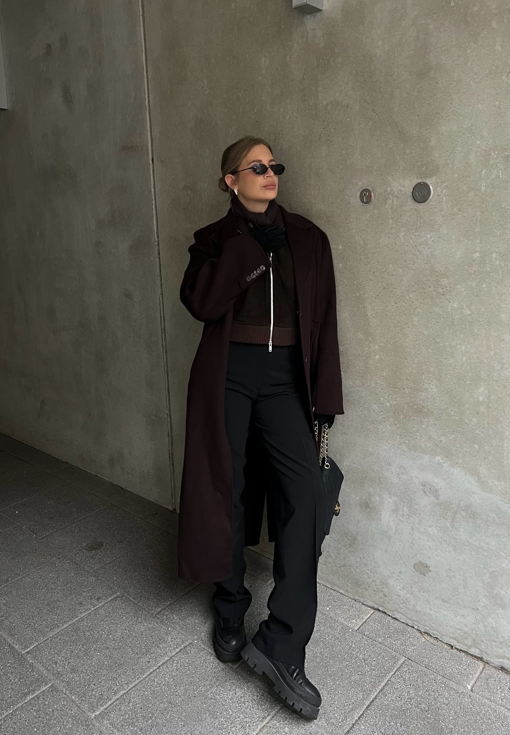 Σκούρο καφέ μακρύ παλτό πάνω από ένα καφέ πουλόβερ με φερμουάρ, μαύρο παντελόνι σε φαρδιά γραμμή και χοντρές μαύρες μπότες. Τα αξεσουάρ περιλαμβάνουν μικρά γυαλιά ηλίου και μια τσάντα.