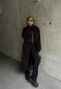 Σκούρο καφέ μακρύ παλτό πάνω από ένα καφέ πουλόβερ με φερμουάρ, μαύρο παντελόνι σε φαρδιά γραμμή και χοντρές μαύρες μπότες. Τα αξεσουάρ περιλαμβάνουν μικρά γυαλιά ηλίου και μια τσάντα.