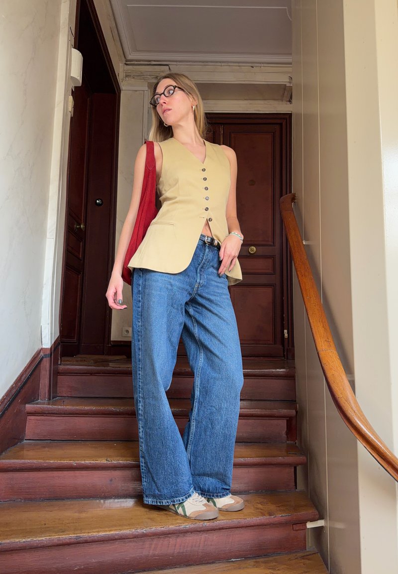Jeune femme portant des lunettes, un gilet sans manches beige boutonné, un jean large bleu, des baskets blanches et un sac rouge, debout sur des escaliers en bois.