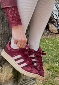 Burgundski Adidas Campus tenisky z usnja s simboli v belem, strukturiranim jezikom in ujemajočimi se vezalkami, položene ob deblo drevesa.