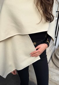 Crèmekleurige wollen mix omslag met een asymmetrisch ontwerp, gelaagde plooien en een gladde textuur; gecombineerd met een zwart topje en riem met gouden details.