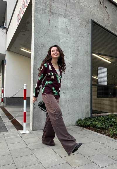 Cardigan burdeos con patrones geométricos verdes, combinado con pantalones anchos marrones. Bolso negro y zapatos negros. Entorno urbano con fondo de concreto.
