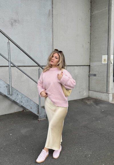 Joven con suéter rosa, falda de satén crema y zapatillas rosas, de pie junto a escaleras metálicas y una pared de hormigón al aire libre.