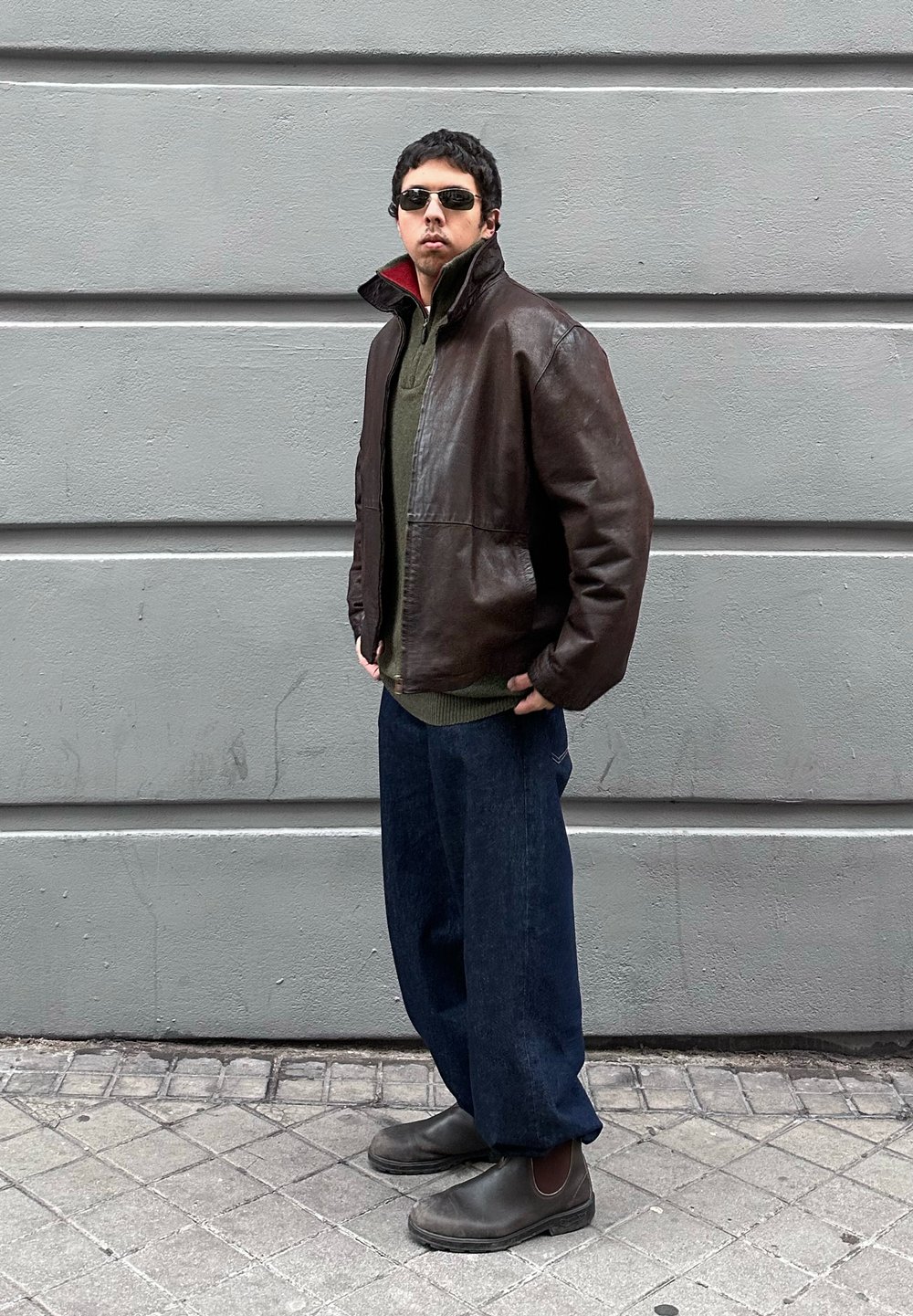 Homme portant des lunettes de soleil noires, une veste en cuir marron, un pull vert, un jean ample foncé et des bottes noires, debout sur un trottoir carrelé.