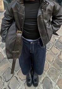 Jachetă din piele neagră, bluză neagră ajustată, pantaloni largi din denim închisi, cizme negre și o geantă de umăr texturată maro. Fundal din piatră pavată.