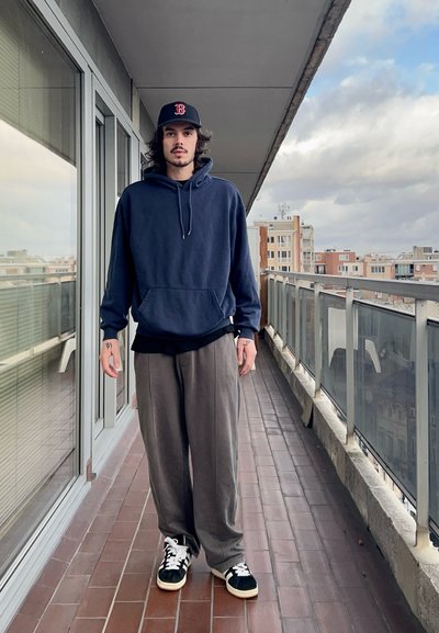 Sudadera navy con bolsillo frontal, pantalones grises de pierna ancha y zapatillas negras con suelas blancas, de pie en un balcón con vista a la ciudad.
