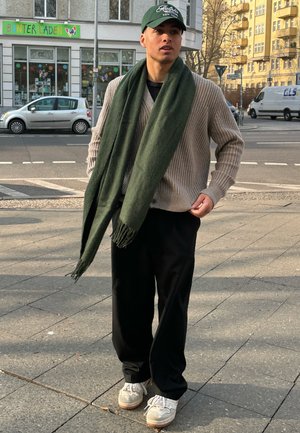 Bufanda verde, cárdigan beige de canalé, pantalones anchos negros y zapatillas grises. El fondo incluye una calle concurrida con edificios y un coche aparcado.