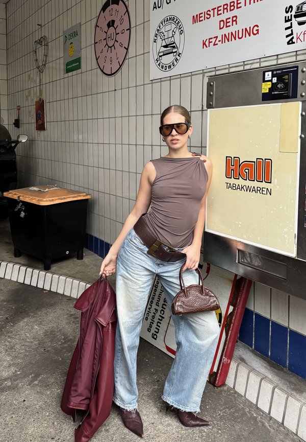 Bruine mouwloze top, hooggewaisted lichtblauwe jeans, bordeauxleren jas, donkerbruine handtas, oversized zonnebril, stedelijke achtergrond.