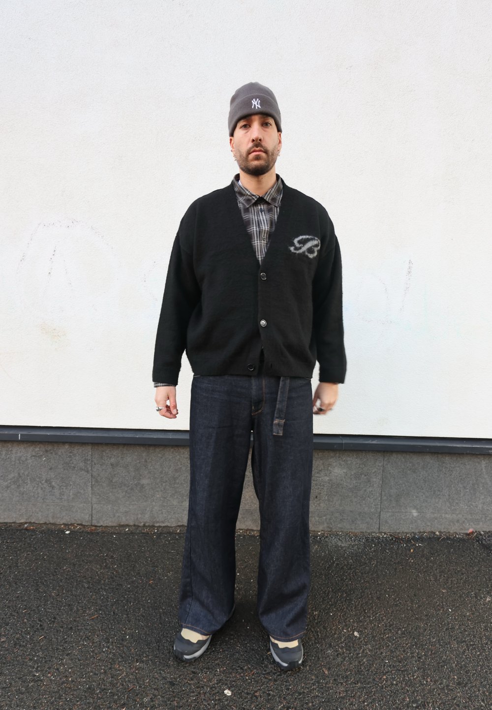 Cardigan noir avec boutons et détail lettre, superposé à une chemise à carreaux. Pantalon en denim foncé ample et bonnet gris complètent le look.