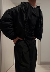 Jachetă bomber neagră cu textură matlasată, purtată peste o cămașă neagră ajustată și pantaloni negri croiți. O brățară argintie adaugă un accent metalic.