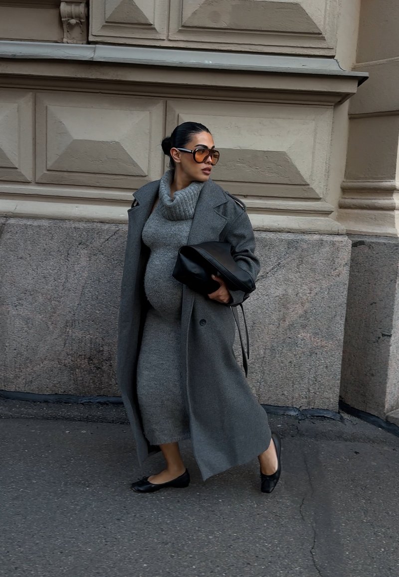 Pregnant woman walking on sidewalk wearing gray knit dress, long gray coat, black flats, large sunglasses, and holding a black clutch bag.