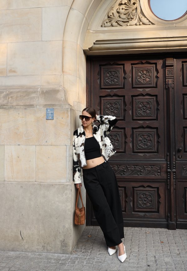 Mujer con chaqueta blanca y negra, top corto negro y pantalones anchos apoyada en una pared de piedra junto a una gran puerta de madera oscura tallada.