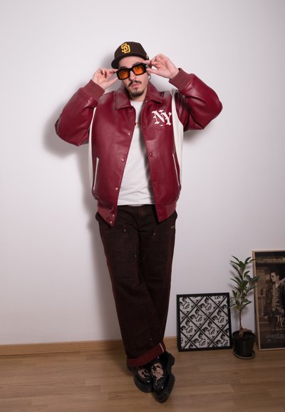 Chaqueta bomber de cuero rojo con acentos blancos y bordado "NY", combinada con pantalones marrón oscuro y calcetines con estampado. Gorra marrón y gafas de sol naranjas.