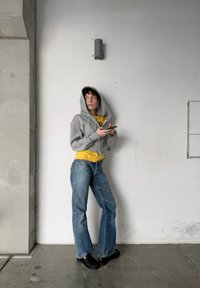 Sweat-shirt à capuche gris, chemise jaune, jeans évasés bleus et chaussures noires. Debout contre un mur blanc avec un luminaire au-dessus.