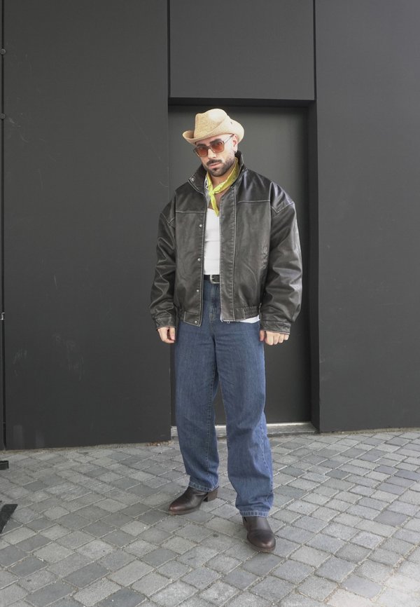 Musta nahkatakki, valkoinen paita, keltainen nauha, siniset farkut. Ruskeat saappaat, olki cowboy-hattu, aurinkolasit. Seisoo tummaa seinää vasten.