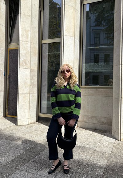 Camisa de rayas de manga larga en verde y azul marino, pantalones de mezclilla oscuros acampanados, bolso negro, zapatos negros, de pie en un entorno urbano.