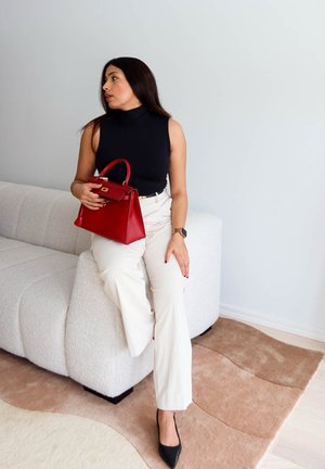 Mujer con top negro sin mangas y pantalones blancos sentada en un sofá blanco, sosteniendo un bolso rojo, mirando de lado en el interior sobre una alfombra beige.