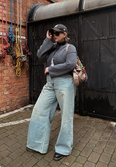 Cárdigan de lana gris, suéter de cuello alto negro, jeans de pierna ancha azul claro, botas negras de plataforma, mochila beige con acentos coloridos, gorra negra y gafas de sol.