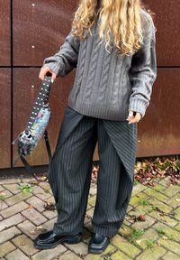 Pull gris surdimensionné en maille torsadée associé à un pantalon noir à fines rayures avec un design drapé unique. Porte un sac à bandoulière multicolore.