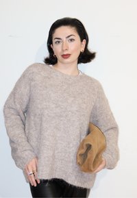 Femme aux cheveux courts et foncés, portant un pull beige et un pantalon noir, tenant une pochette souple marron sur un fond blanc.