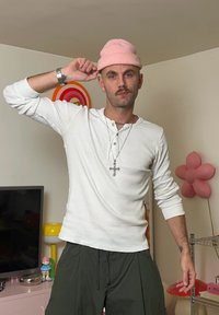 Biela henley košeľa s dlhým rukávom, ružová pletená čiapka, olivovo zelené nohavice, strieborné hodinky a náhrdelník s krížom. Pozadie tvorí farebný dizajn steny.