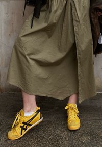 Κίτρινες αθλητικές παπούτσια με μαύρες λεπτομέρειες, διαθέτοντας καπάκι από σουέντ στη μύτη και αντίθετα κορδόνια, συνδυασμένα με μια μακριά φούστα σε ελαιόλευκο χρώμα.