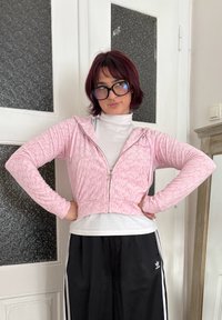 Lühendatud roosa tõmblekaga kapuutsiga sviiter, millel on tekstuuriline muster, kapuuts ja pikad varrukad, seljas valge kõrge kaelusega särk ning kaasas mustad spordipüksid.