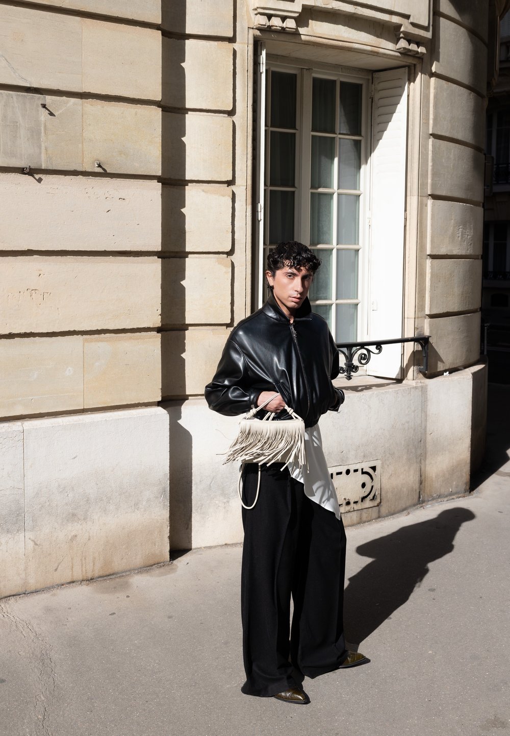 Zwarte leren bomberjack, losse zwarte broek, witte asymmetrische shirt en een franjes handtas in crèmekleur. Zonnige stedelijke omgeving met een beige stenen muur.