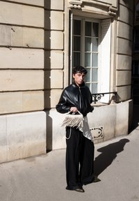 Zwarte leren bomberjack, losse zwarte broek, witte asymmetrische shirt en een franjes handtas in crèmekleur. Zonnige stedelijke omgeving met een beige stenen muur.