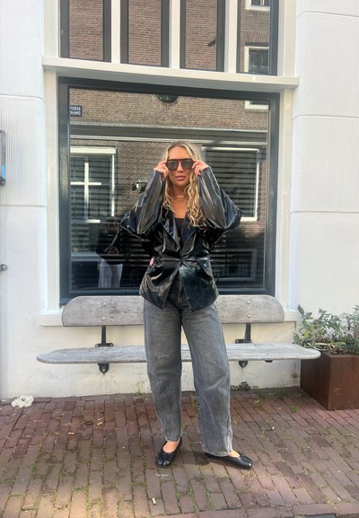 Chaqueta de charol negro con mangas oversized, combinada con pantalones de pierna ancha gris y zapatos planos negros. La modelo lleva gafas de sol oscuras.