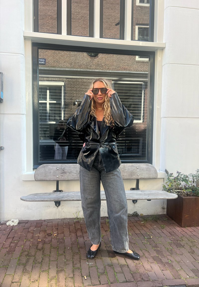 Giacca in pelle verniciata nera con maniche oversize, abbinata a jeans grigi a gamba larga e ballerine nere. Il modello indossa occhiali da sole scuri.