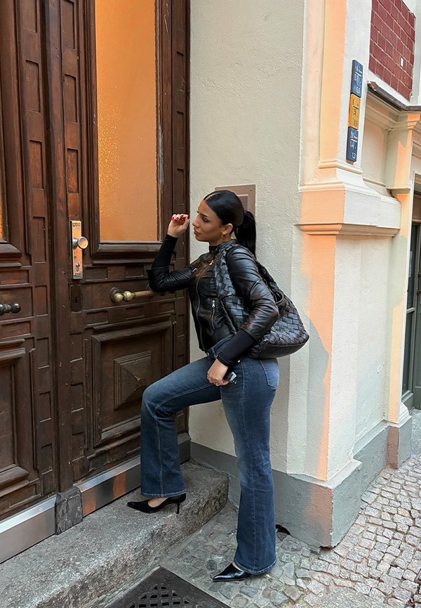 Black leather jacket, dark flared jeans, black high heels, woven black handbag. Standing at a wooden door with warm lighting.