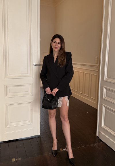 Mujer con un vestido blazer negro con ribete de encaje blanco sostiene un bolso tejido negro, de pie en una habitación beige con suelos de madera.