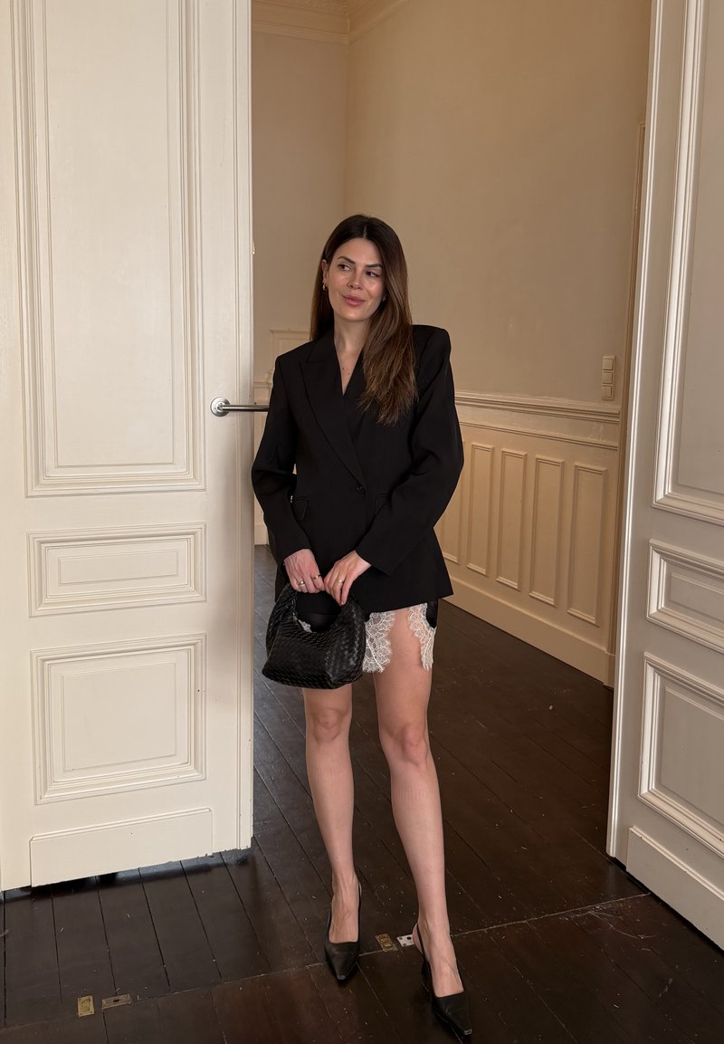 Donna con un abito blazer nero con bordi in pizzo bianco tiene in mano una borsa nera intrecciata, in piedi in una stanza beige con pavimenti in legno.