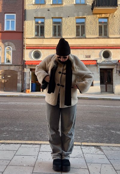Chaqueta de forro polar crema con mangas amplias, bufanda negra y gorro negro, combinada con pantalones grises y zapatos de plataforma, al aire libre con un fondo de ciudad.