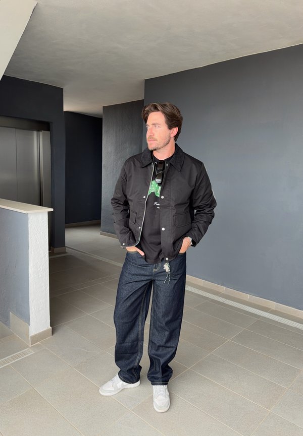 Homme en veste décontractée sombre, t-shirt noir, jean bleu et baskets blanches debout dans un couloir près d'un ascenseur, regardant sur le côté.