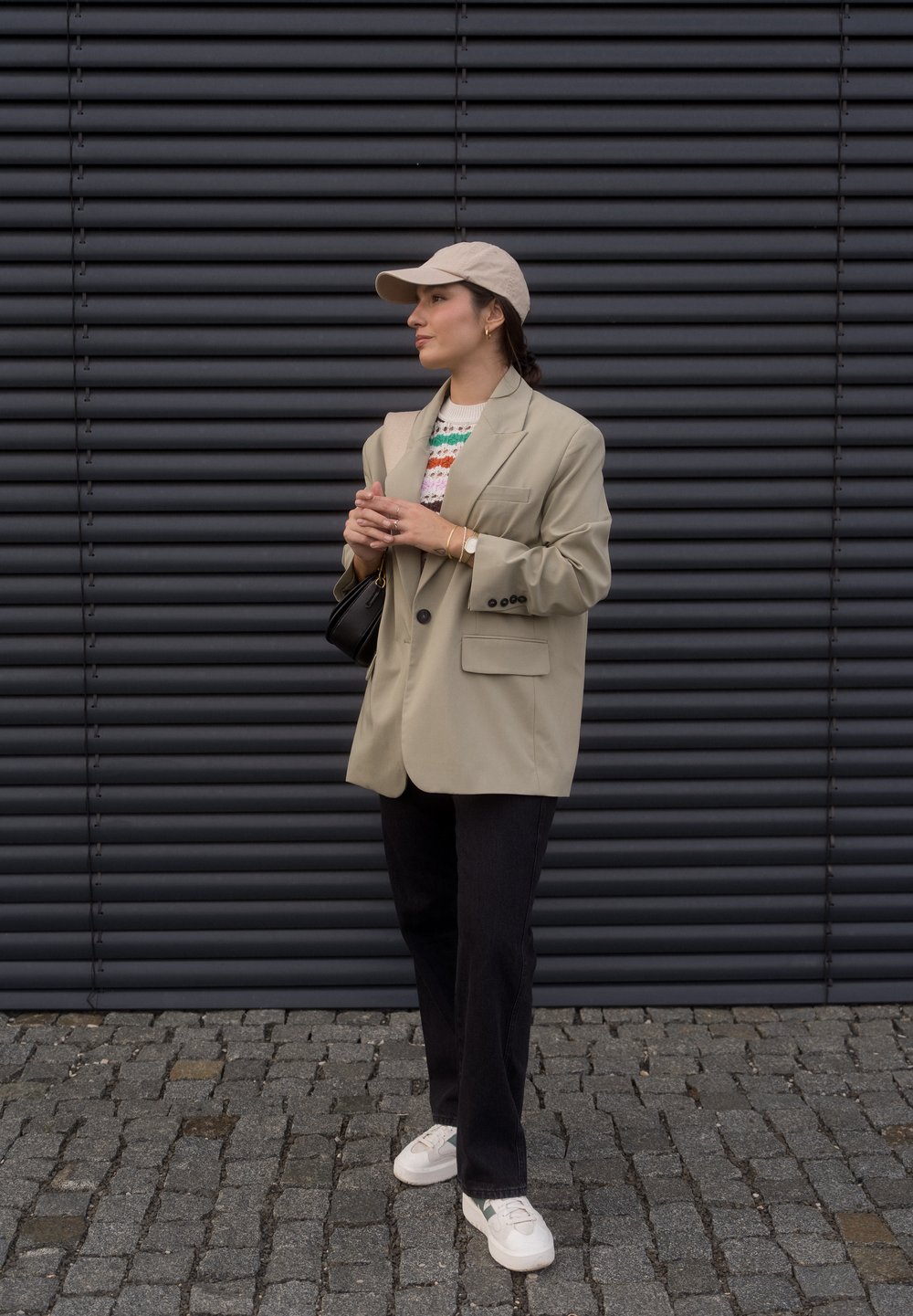 Blazer oversized bege sobre um suéter de malha multicolorido, combinado com jeans pretos e ténis brancos. Um boné claro e uma pequena bolsa completam o visual.