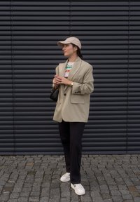 Blazer oversized bege sobre um suéter de malha multicolorido, combinado com jeans pretos e ténis brancos. Um boné claro e uma pequena bolsa completam o visual.