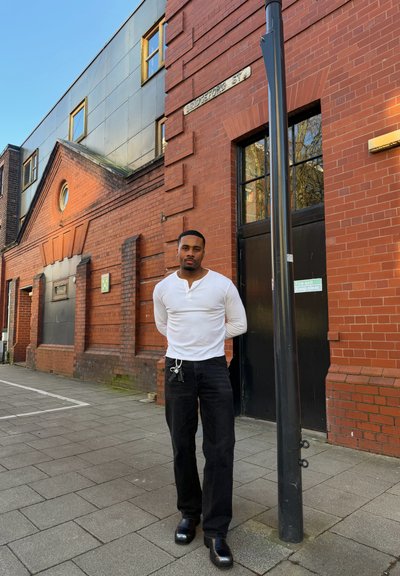 Hombre con camisa blanca de manga larga y pantalones negros de pie en la acera de Bridgeford Street cerca de un edificio de ladrillo rojo y una puerta negra con señal de salida de emergencia.