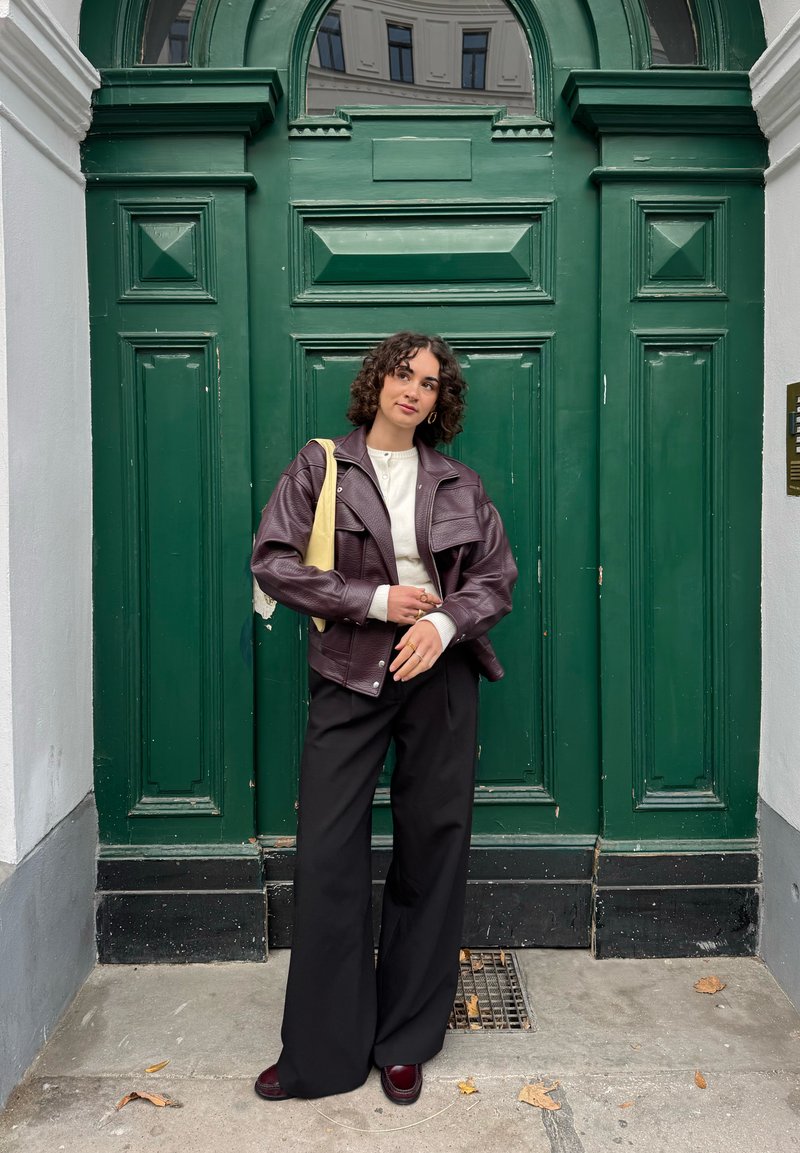 Bordeauxrotes Lederjacke mit zwei Brusttaschen, cremefarbener Pullover, weite schwarze Hose und bordeauxrote Schuhe vor einer dunkelgrünen Türkulisse.