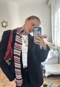 Black blazer worn over a white top, decorated with a multicoloured striped scarf. Gold layered necklaces enhance the look, paired with a burgundy handbag.
